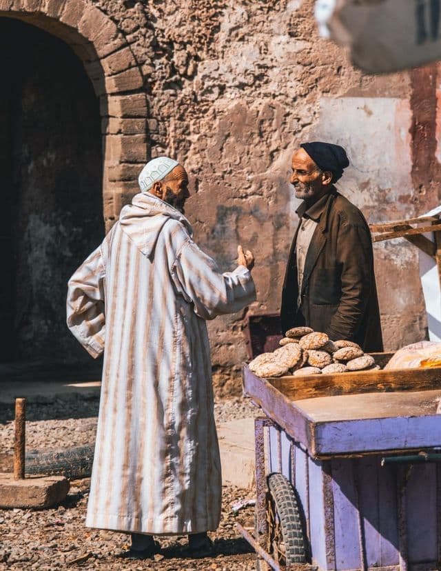 Due uomini in abiti tradizionali parlano accanto a un carretto viola che vende focacce, davanti a un muro di pietra rustico.