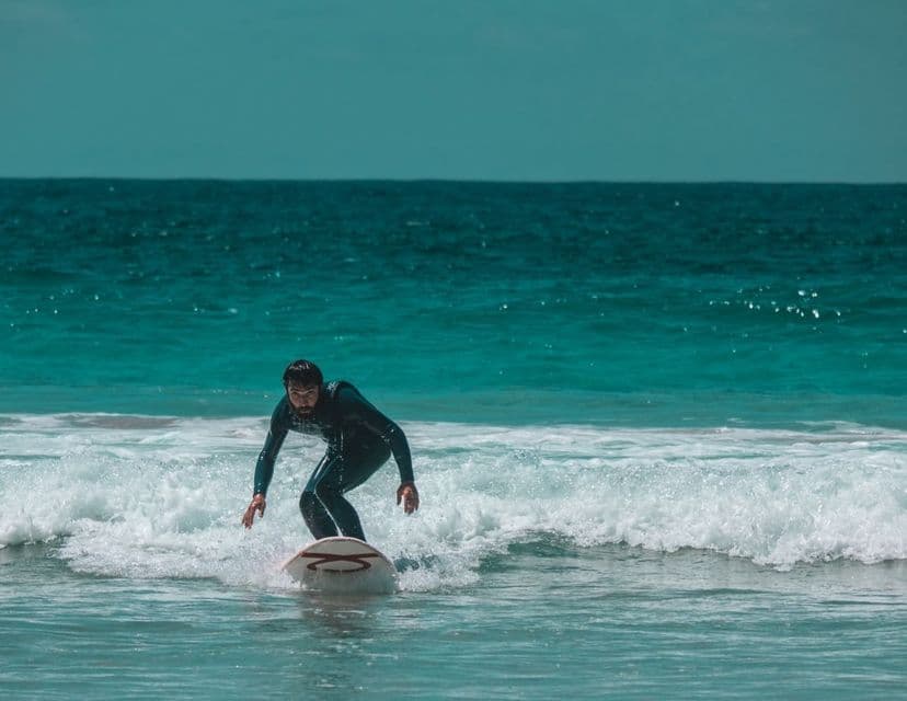 Un uomo in muta nera si accovaccia sulla sua tavola da surf, cavalcando una piccola onda nell'acqua turchese dell'oceano.