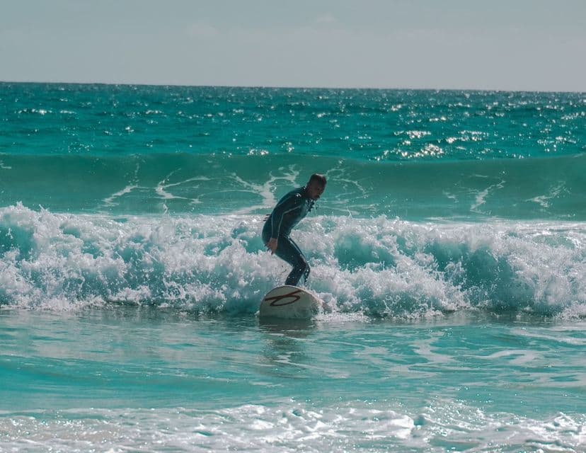 Una persona in muta nera sta su una tavola da surf, cavalcando un'onda che si infrange in acqua turchese brillante.