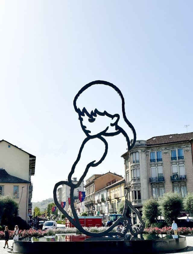 Una grande scultura a sagoma nera di una ragazza è seduta in una fontana in una vivace piazza cittadina sotto il sole.