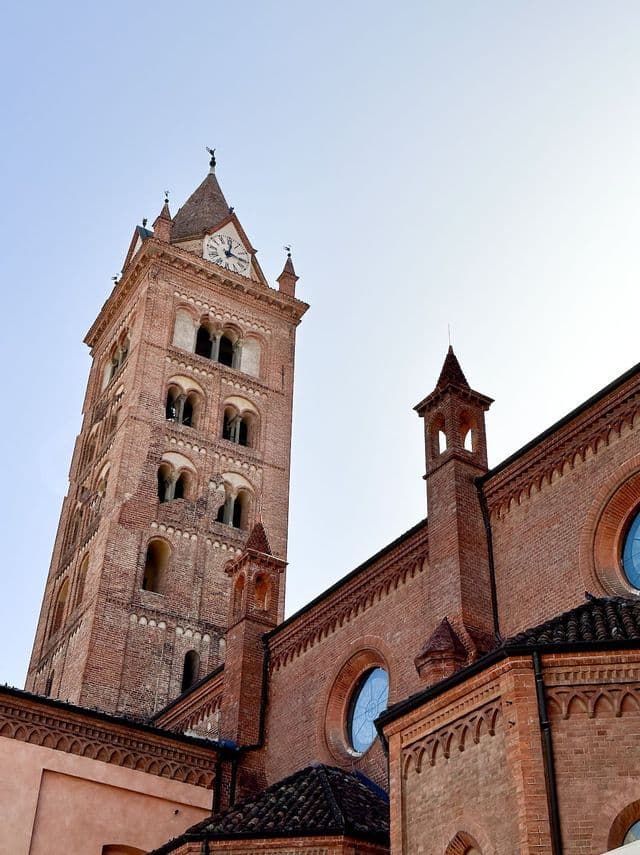 Una vista dal basso di un'alta torre dell'orologio in mattoni rossi, accanto a una chiesa, su sfondo di cielo azzurro limpido.