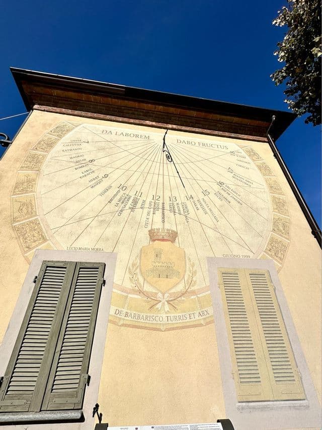 Una grande meridiana ornata, dipinta sulla parete esterna di un edificio, ripresa dal basso, con due finestre con persiane sottostanti, contro un cielo azzurro e limpido.