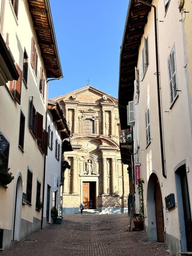 Una vista su un vicolo stretto e acciottolato, incorniciato da edifici, che porta all'ingresso di un'ornata chiesa in mattoni sotto un cielo azzurro.