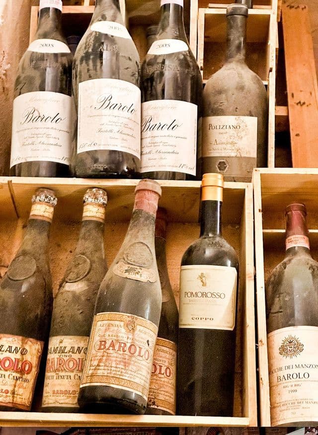 Una collezione di vecchie bottiglie impolverate di vino Barolo che riposano su scaffali di legno in una cantina.