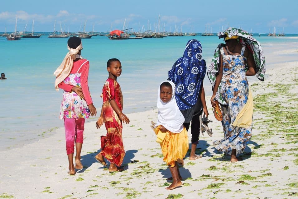 Un groupe de femmes et d'enfants vêtus de vêtements colorés marche le long d'une plage de sable blanc, avec une eau turquoise et des bateaux en arrière-plan.