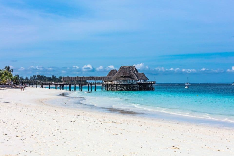 Ein Holzgebäude mit Reetdach auf Stelzen steht am Ende eines Stegs über türkisfarbenem Wasser, neben einem weißen Sandstrand.