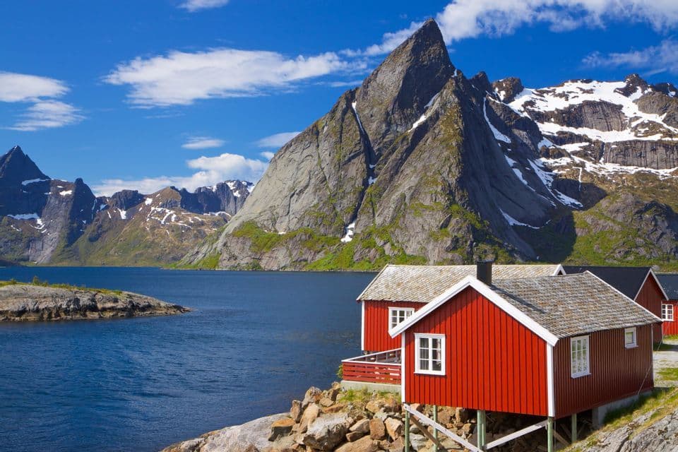Red wooden cabins on stilts sit on a rocky shore beside blue water, with steep, snow-capped mountains under a clear sky.