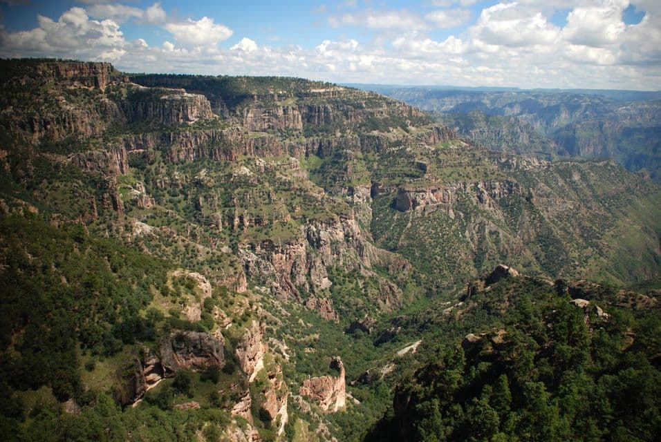 Una vista panoramica su un vasto sistema di canyon con ripide scogliere rocciose e pendii ricoperti di alberi e arbusti verdi sotto un cielo blu, parzialmente nuvoloso.