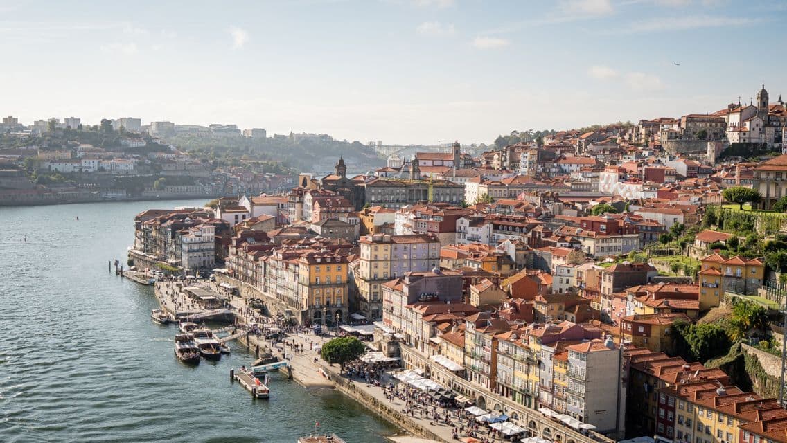 A high-angle view of a city with colorful buildings and terracotta roofs built on a hillside next to a wide river.