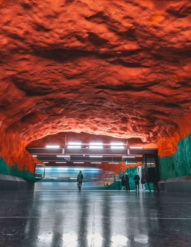 Un train traverse une station de métro avec un plafond rouge évoquant une grotte et des murs peints, alors que des passagers attendent sur le quai réfléchissant.