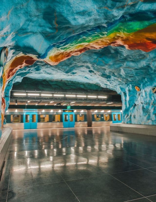 Un treno della metropolitana su una banchina con un soffitto a forma di grotta dipinto di blu e una striscia arcobaleno colorata.