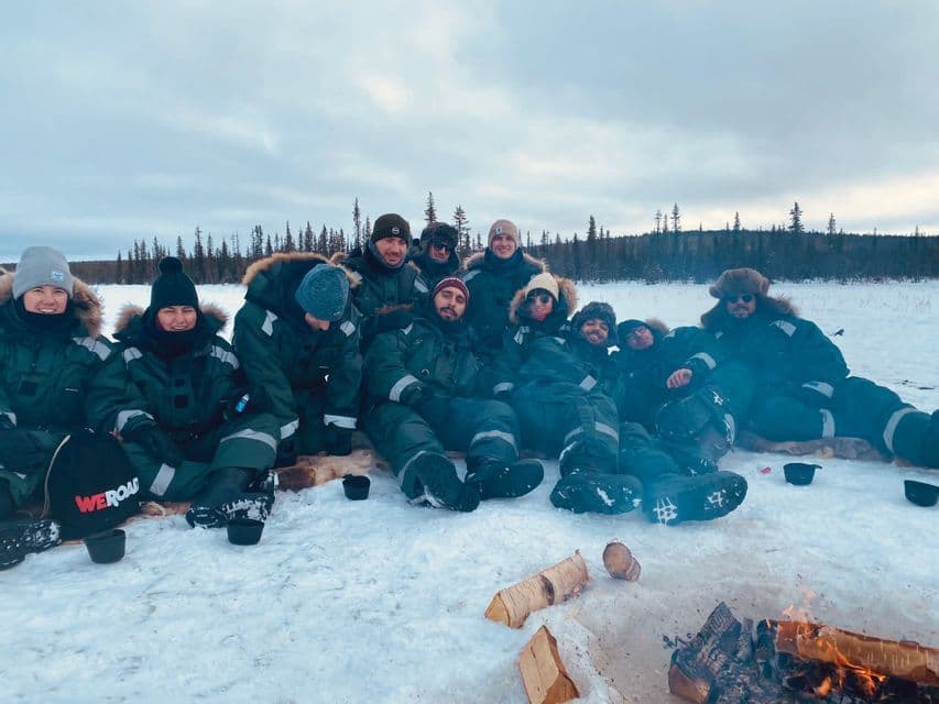 Un groupe WeRoad réuni dans la neige autour d'un petit feu de camp, avec une forêt en arrière-plan.