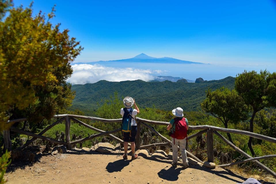 Zwei Wanderer mit Rucksäcken stehen an einem Aussichtspunkt und blicken auf einen Berggipfel, der über den Wolken über einem grünen Tal emporragt.