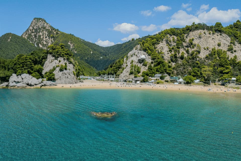 Una vista panoramica di una caletta sabbiosa con acqua turchese, circondata da montagne verdi sotto un cielo azzurro e soleggiato.