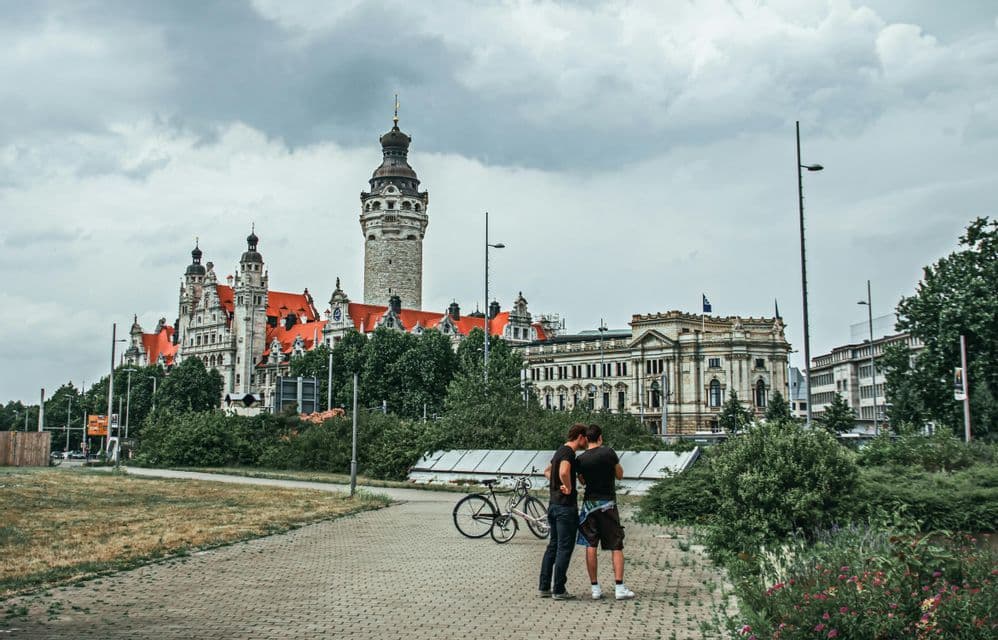 Zwei Personen stehen mit einem Fahrrad auf einem gepflasterten Weg, im Hintergrund große historische Gebäude und ein Steinturm.