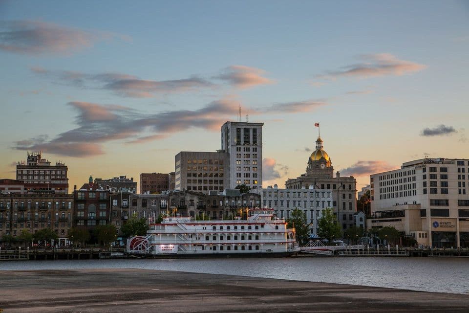 The "Georgia Queen" riverboat is docked on a river in front of a city skyline with a golden-domed building at sunset.