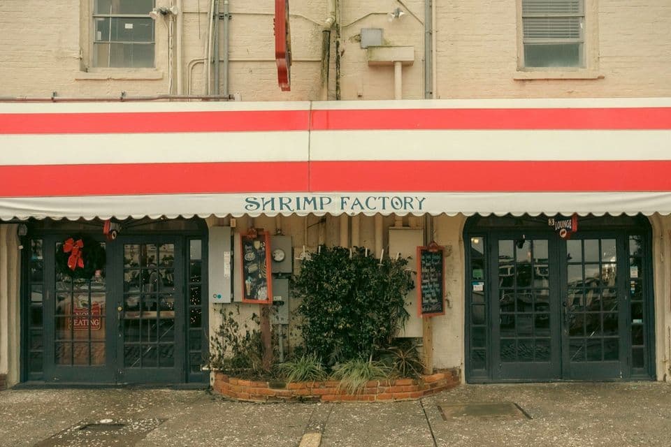 The storefront of the 'Shrimp Factory' restaurant, featuring a red and white striped awning over two glass-paned doors on a brick building.