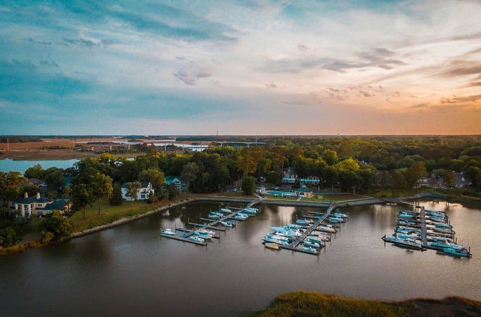An aerial view of a marina with boats docked along piers, with a tree-lined shore and houses under a sunset sky.