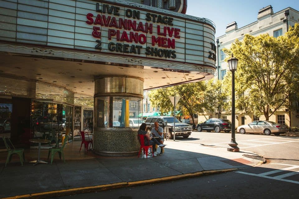 The entrance of a retro-style theater with a large marquee advertising 'Savannah Live & Piano Men' on a sunny sidewalk.