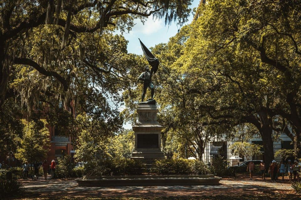 A bronze statue of a soldier holding a flag on a pedestal in a park, surrounded by large trees with Spanish moss.