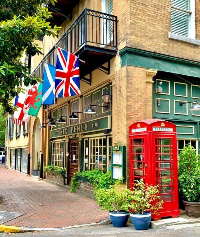 A classic red telephone booth stands on a brick sidewalk outside a British-style pub decorated with UK flags.