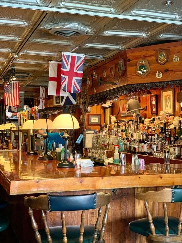 Interior view of a traditional pub with a long wooden bar, stools, and various national flags hanging from the tin ceiling.