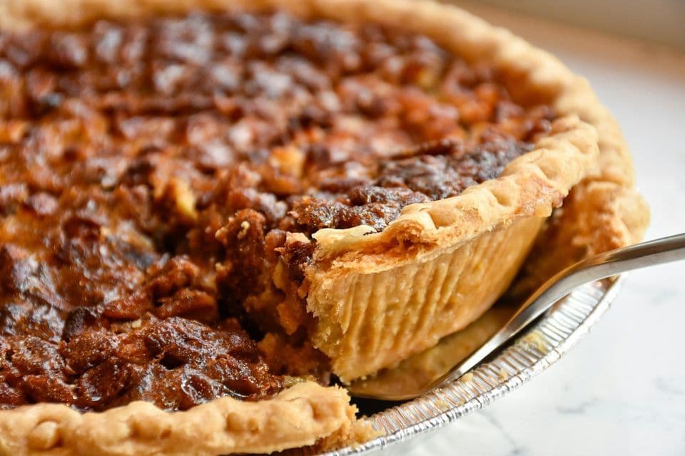 A close-up of a fork lifting a slice from a pecan pie with a flaky crust in a foil pan.