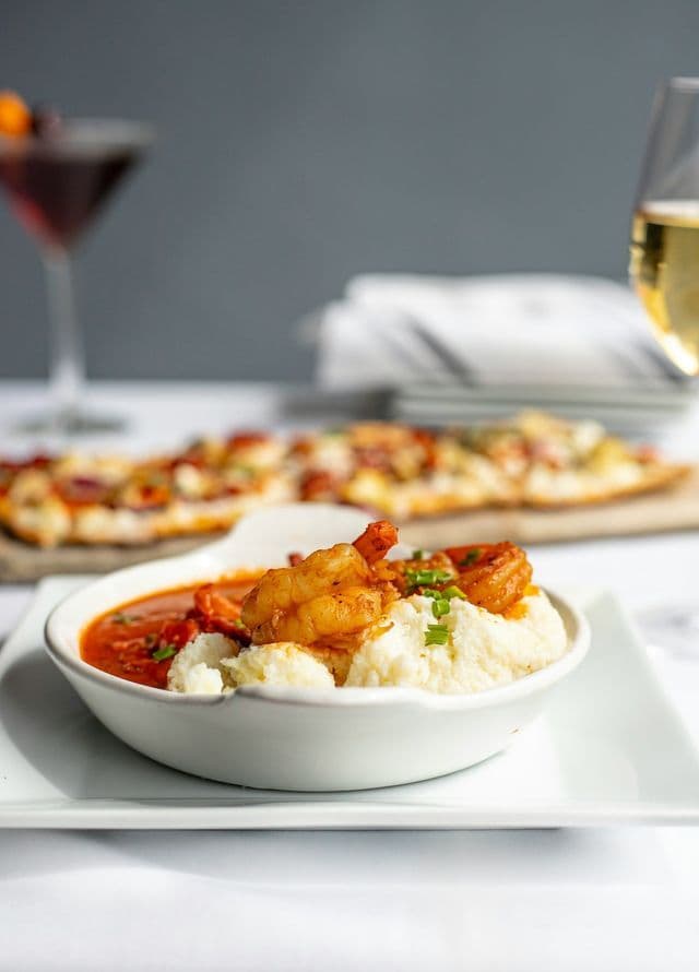 A close-up of a white bowl of shrimp and grits, with a flatbread pizza and drinks blurred in the background.