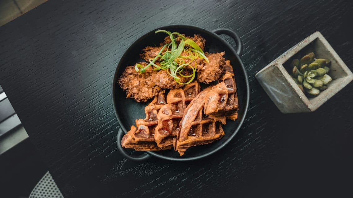 An overhead view of a black bowl containing fried chicken and waffles, garnished with green onions, on a dark table.