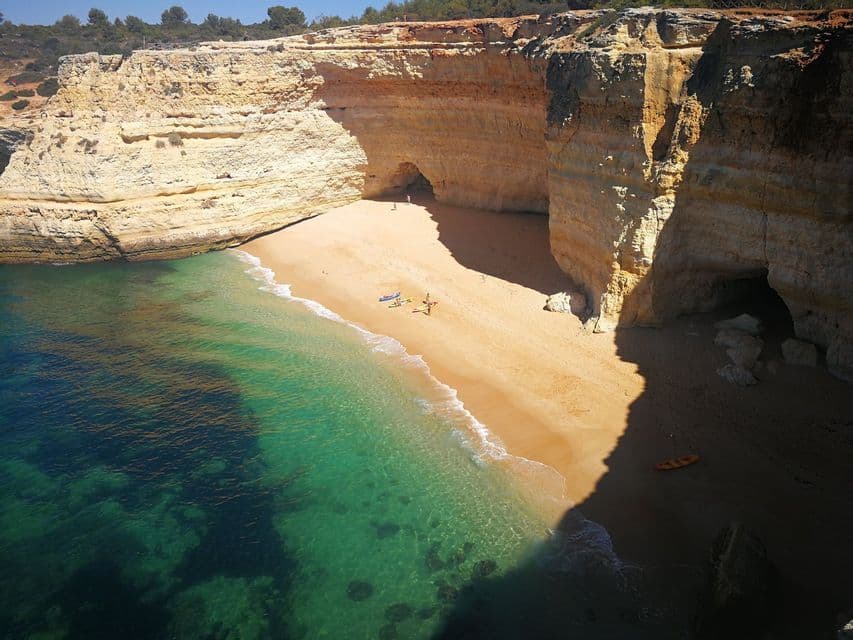 Una spiaggia sabbiosa appartata con acqua cristallina color turchese è circondata da alte scogliere con grotte, vista dall'alto.