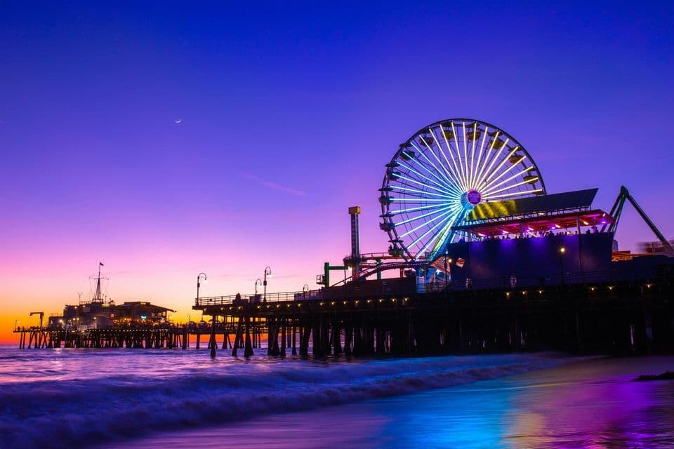An illuminated Ferris wheel on an amusement pier glows against a purple and orange twilight sky over the ocean.