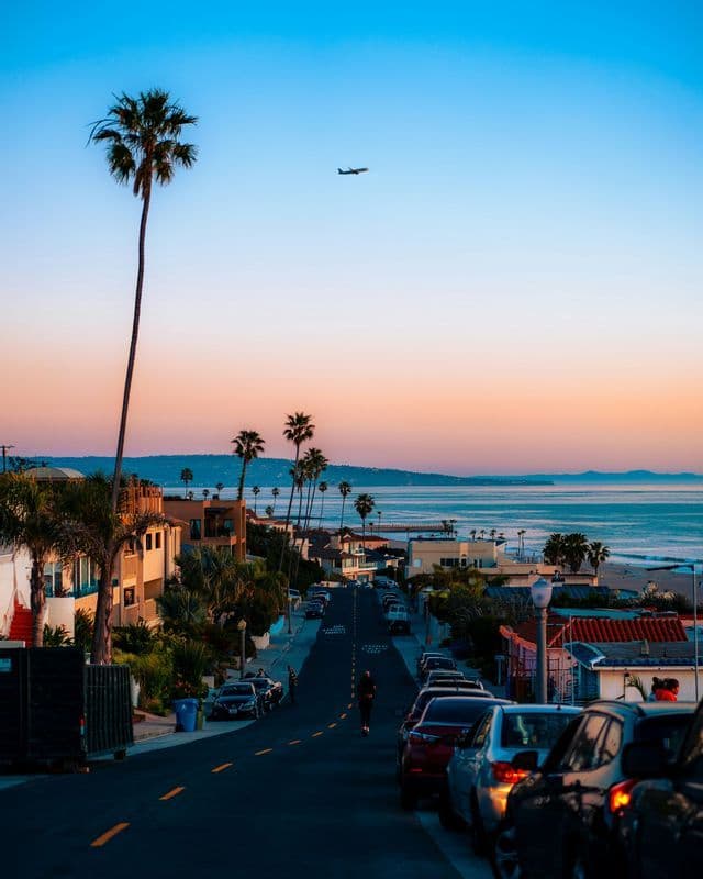 A street lined with palm trees and parked cars slopes down towards the ocean at sunset, as an airplane flies overhead.