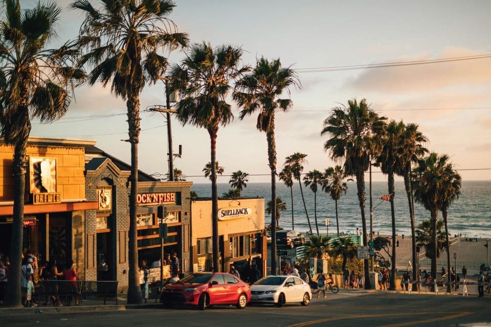 A street lined with palm trees and shops slopes down towards a sandy beach and the ocean during a warm sunset.
