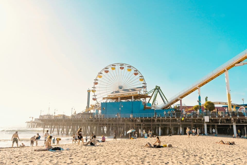 People relax on a sandy beach in front of a pier holding an amusement park with a Ferris wheel and roller coaster.