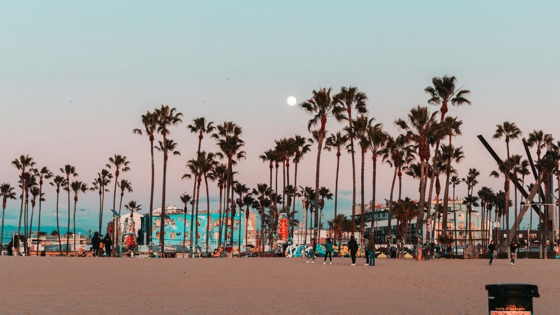A wide sandy beach at dusk with people, silhouetted palm trees, and colorful buildings under a pastel sky with a full moon.
