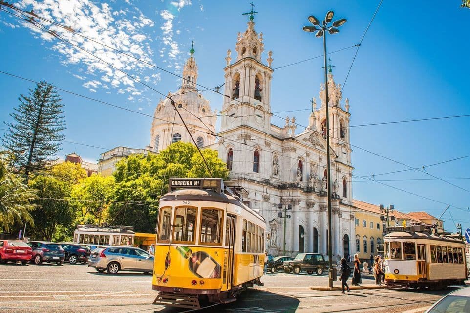 Eine klassische gelbe Straßenbahn auf einer Stadtstraße mit einer verzierten weißen Basilika im Hintergrund unter einem sonnigen blauen Himmel.