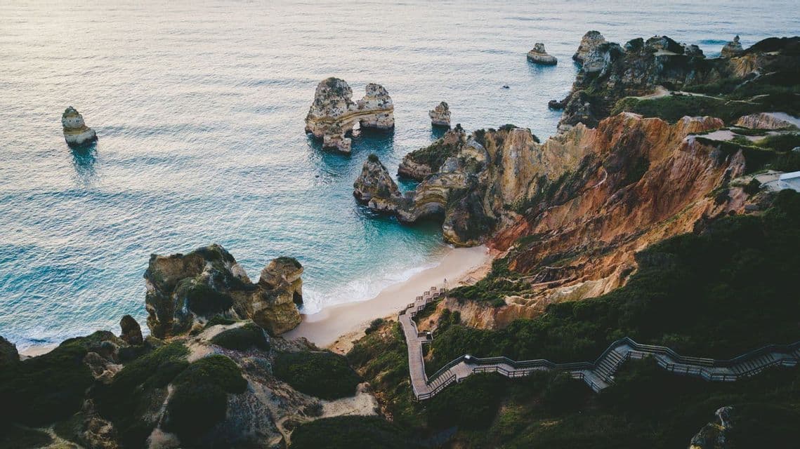 Une vue aérienne d'un escalier en bois serpentant le long d'une falaise rocheuse et verdoyante jusqu'à une plage de sable isolée avec de grands stacks marins dans l'eau.