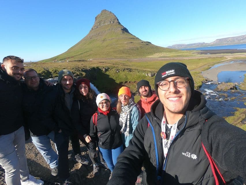 A WeRoad group trip takes a selfie smiling in front of a large green mountain and a river on a sunny day.