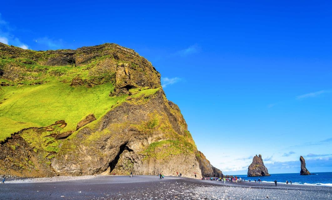 A WeRoad group trip on a black sand beach next to a mossy cliff, with sea stacks visible in the ocean under a clear blue sky.