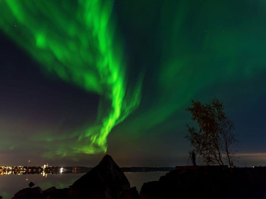The green aurora borealis swirls in the night sky over a body of water, with a photographer silhouetted on the shore.