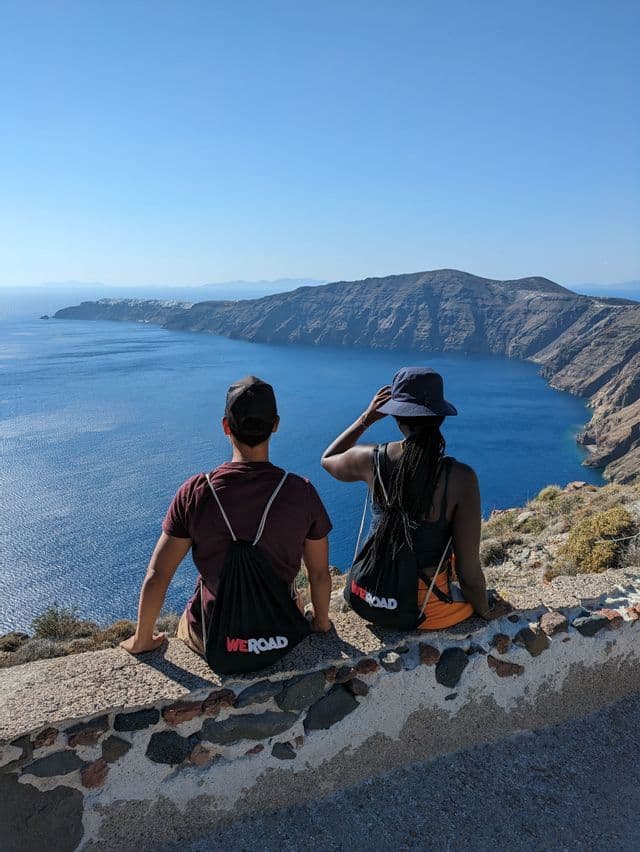 Due persone di un viaggio di gruppo WeRoad sono sedute su un muretto di pietra con vista su una grande baia con scogliere ripide e rocciose sotto un cielo azzurro e limpido.