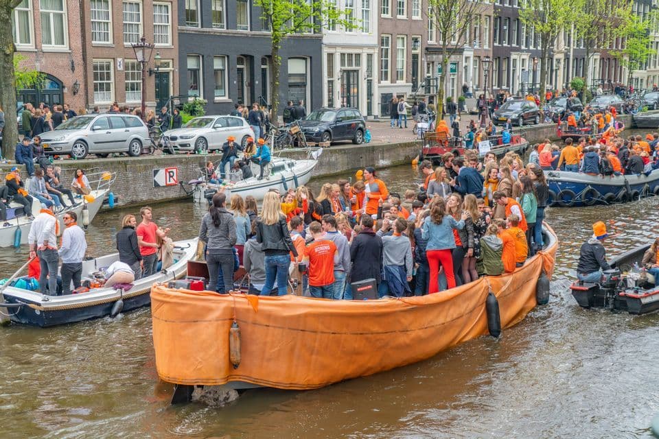 Diverse barche affollate di persone, molte vestite di arancione, navigano lungo un trafficato canale cittadino fiancheggiato da edifici.