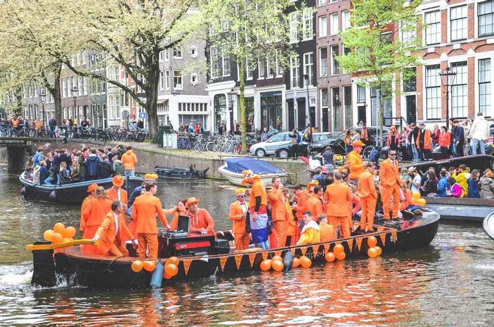 Un grande gruppo di persone che indossano tute arancione brillante festeggia su una barca decorata che naviga lungo un canale cittadino.