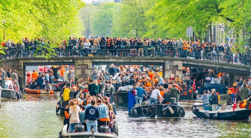 Barche affollate navigano un canale durante un festival, con gente che osserva da un ponte.