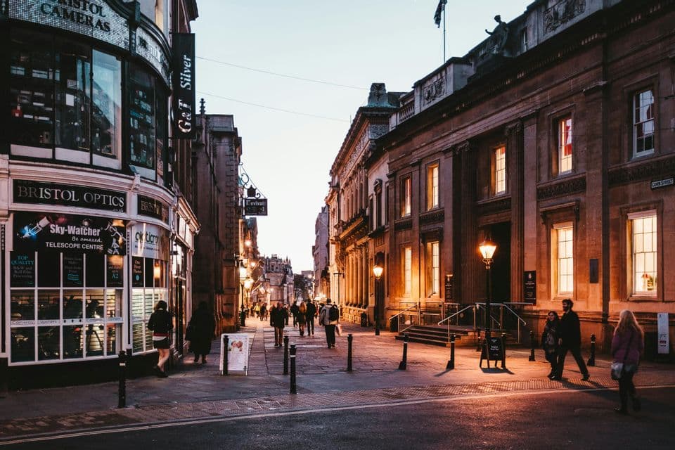 Le persone passeggiano lungo una via storica della città al tramonto, illuminata dalla calda luce dei lampioni e dei negozi.