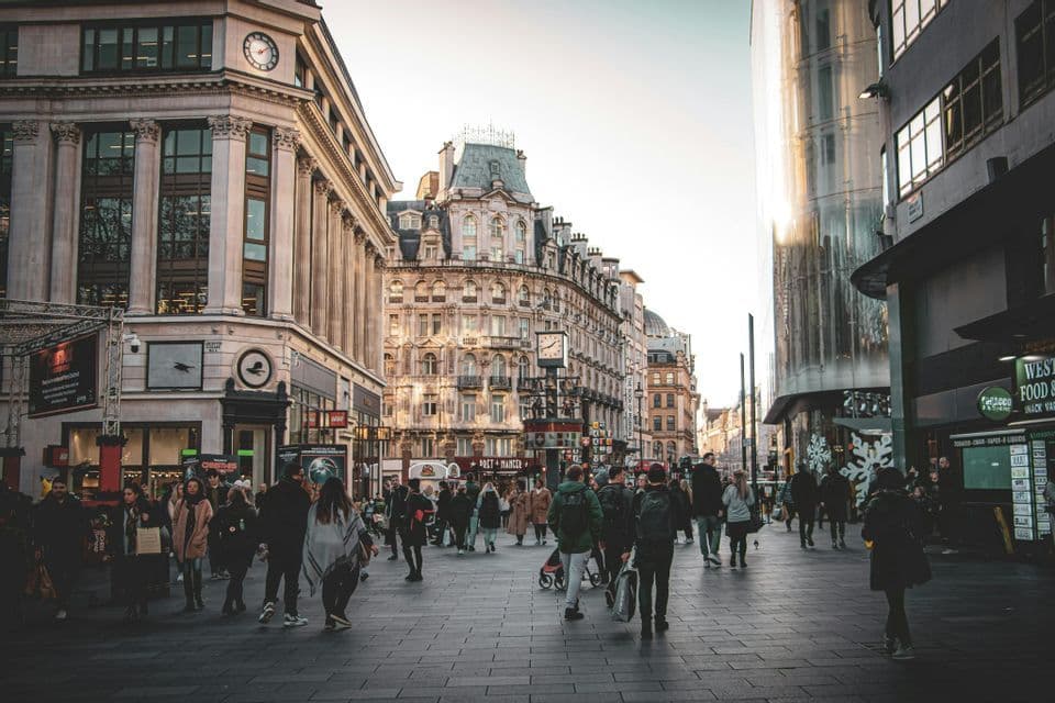 Una folla di persone cammina su un'ampia strada cittadina pavimentata, fiancheggiata da edifici storici ornati e moderni in vetro.