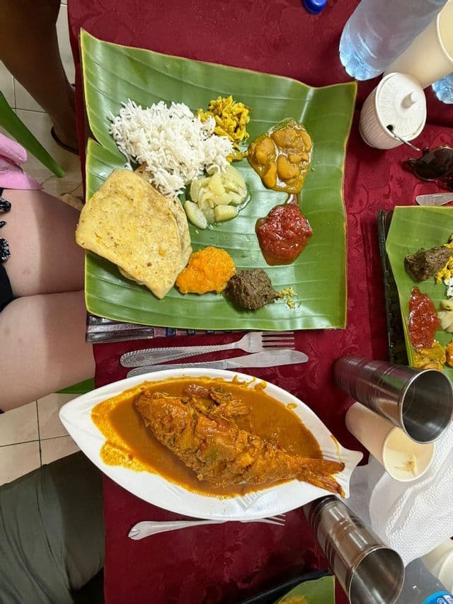 Un repas servi sur une feuille de bananier, composé de riz, de pain plat et de divers accompagnements, à côté d'une assiette blanche contenant un curry de poisson entier.
