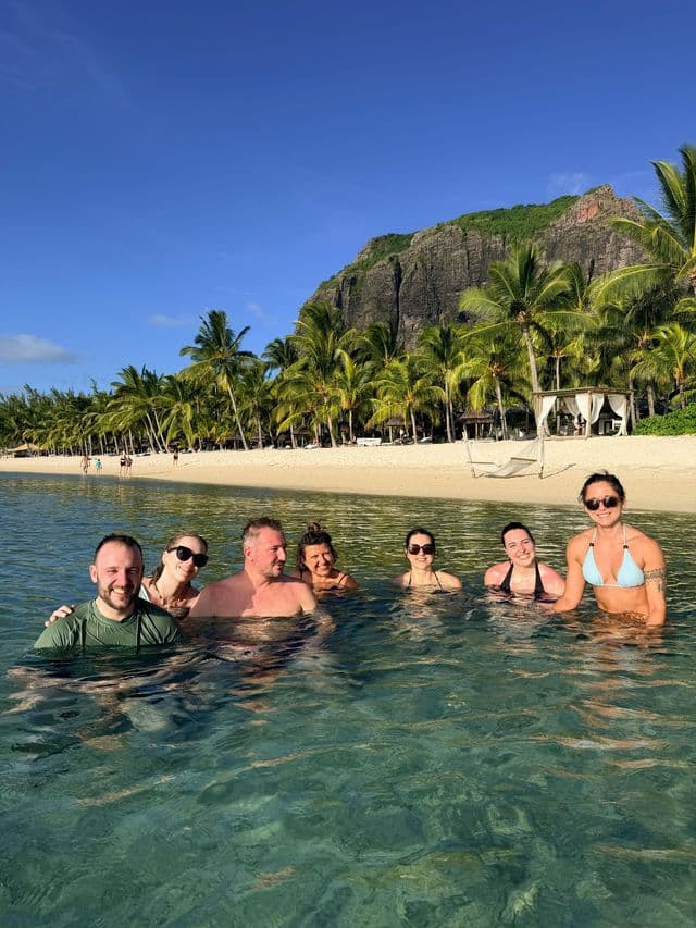 Un voyage de groupe WeRoad souriant dans des eaux tropicales claires, avec une plage de sable, des palmiers et une montagne derrière eux.