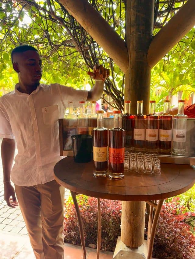 Un homme en chemise blanche présente des bouteilles de rhum et des verres pour une dégustation sur une table en bois en extérieur.