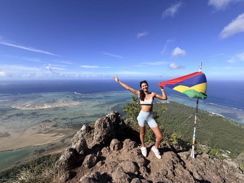 Une femme souriante se tient sur un sommet de montagne rocheux, tenant le drapeau de l'île Maurice, avec l'océan et le littoral en arrière-plan.